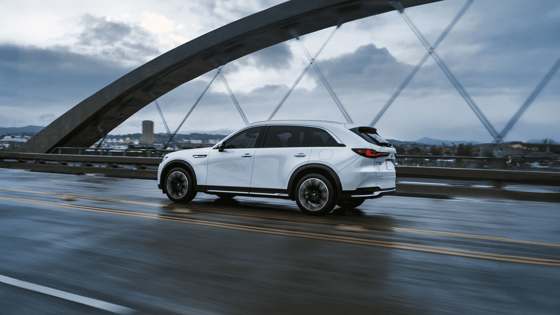 White 2026 Mazda CX-90 driving on a wet bridge in side profile with steel arches, cloudy sky, and distant city skyline