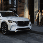 White 2026 Mazda CX-90 parked at a curb outside a brick building, front three-quarter view with two people walking nearby