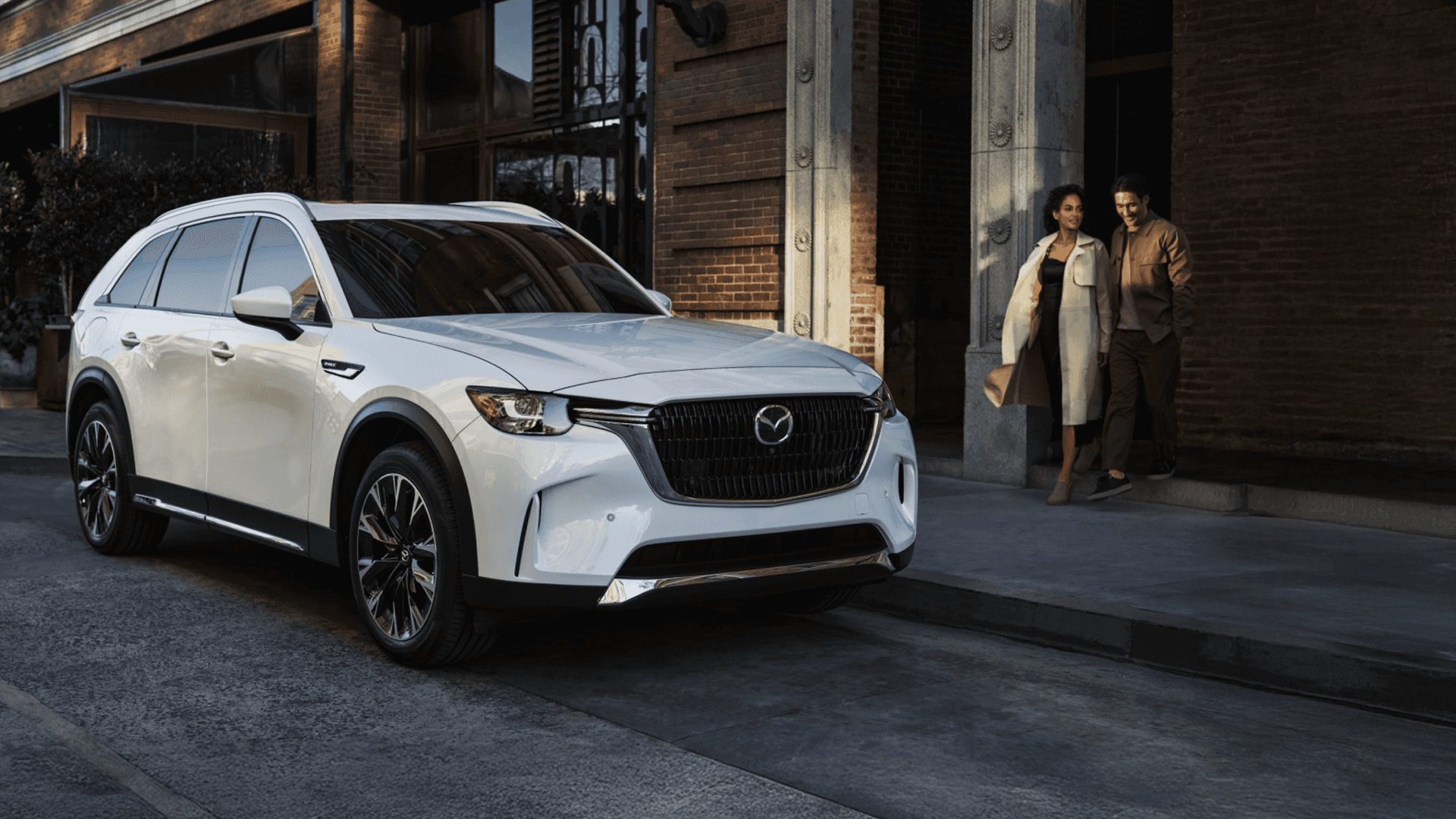 White 2026 Mazda CX-90 parked at a curb outside a brick building, front three-quarter view with two people walking nearby