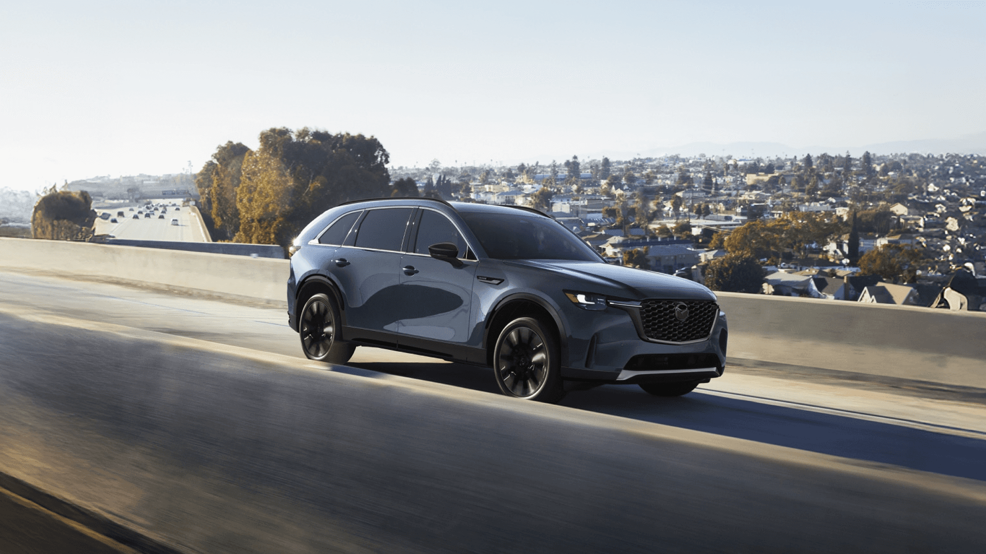 Gray 2026 Mazda CX-90 driving on an elevated roadway, front three-quarter view with black wheels and cityscape background