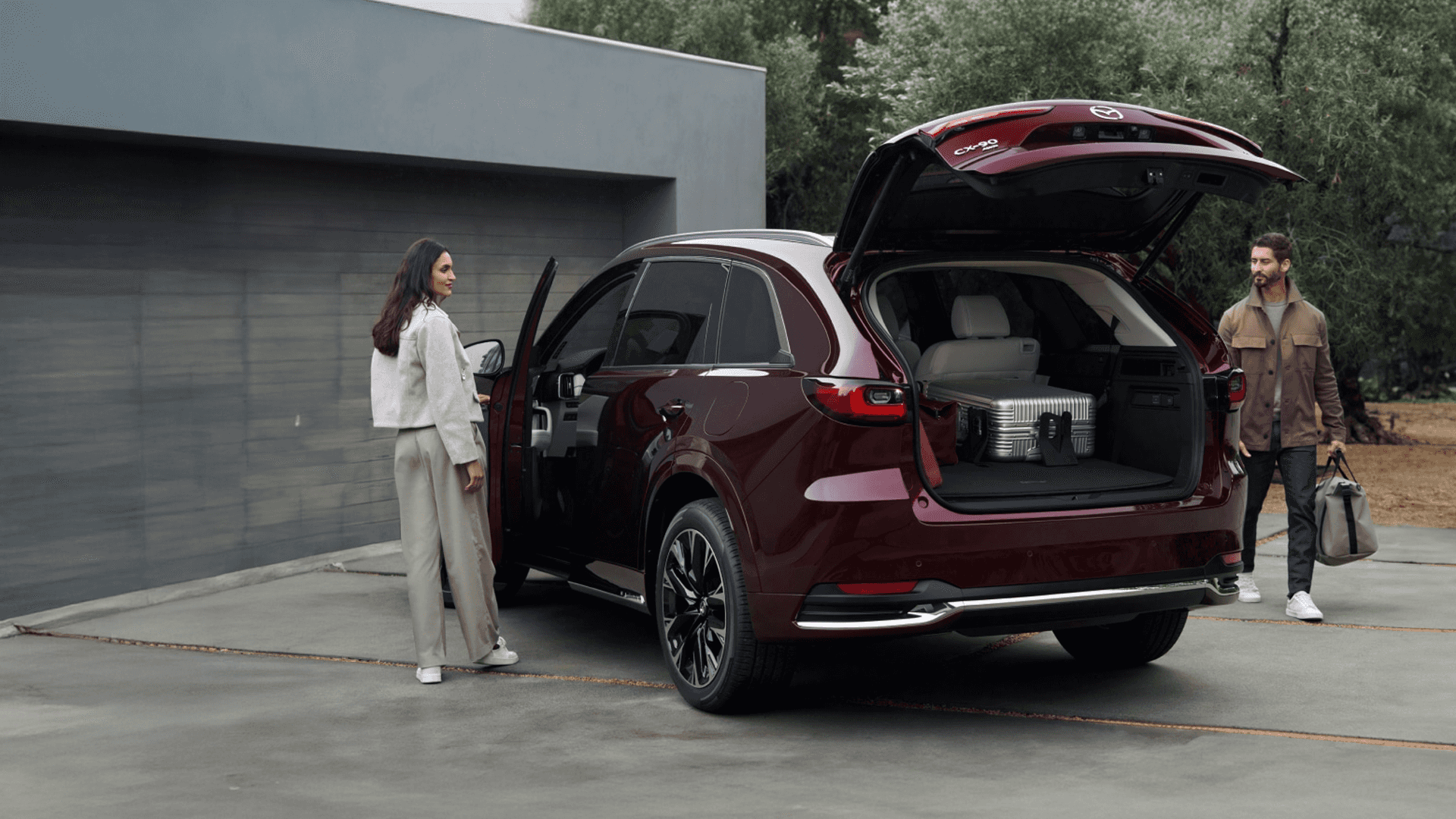 Red 2026 Mazda CX-90 with rear liftgate open showing luggage in cargo area and two people standing beside the vehicle