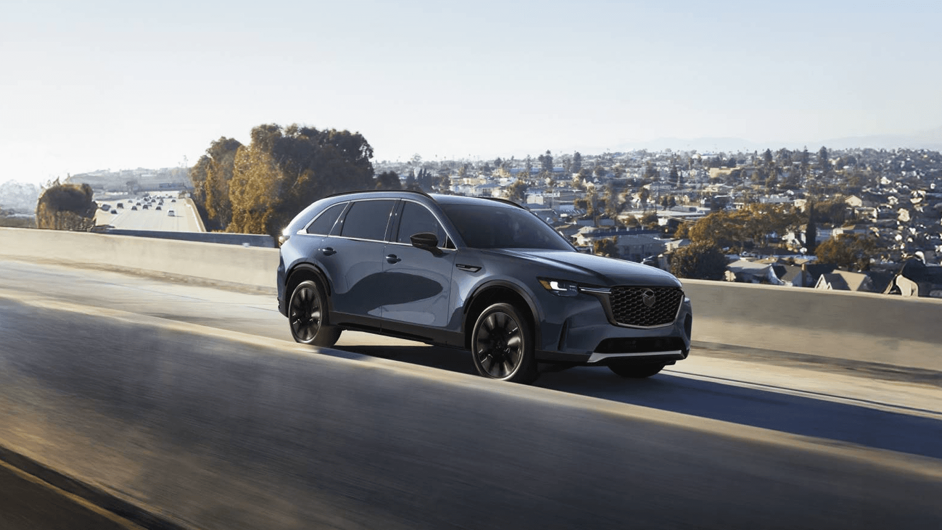 Blue Mazda CX-90 driving on a highway with city skyline and trees in the background under clear daylight