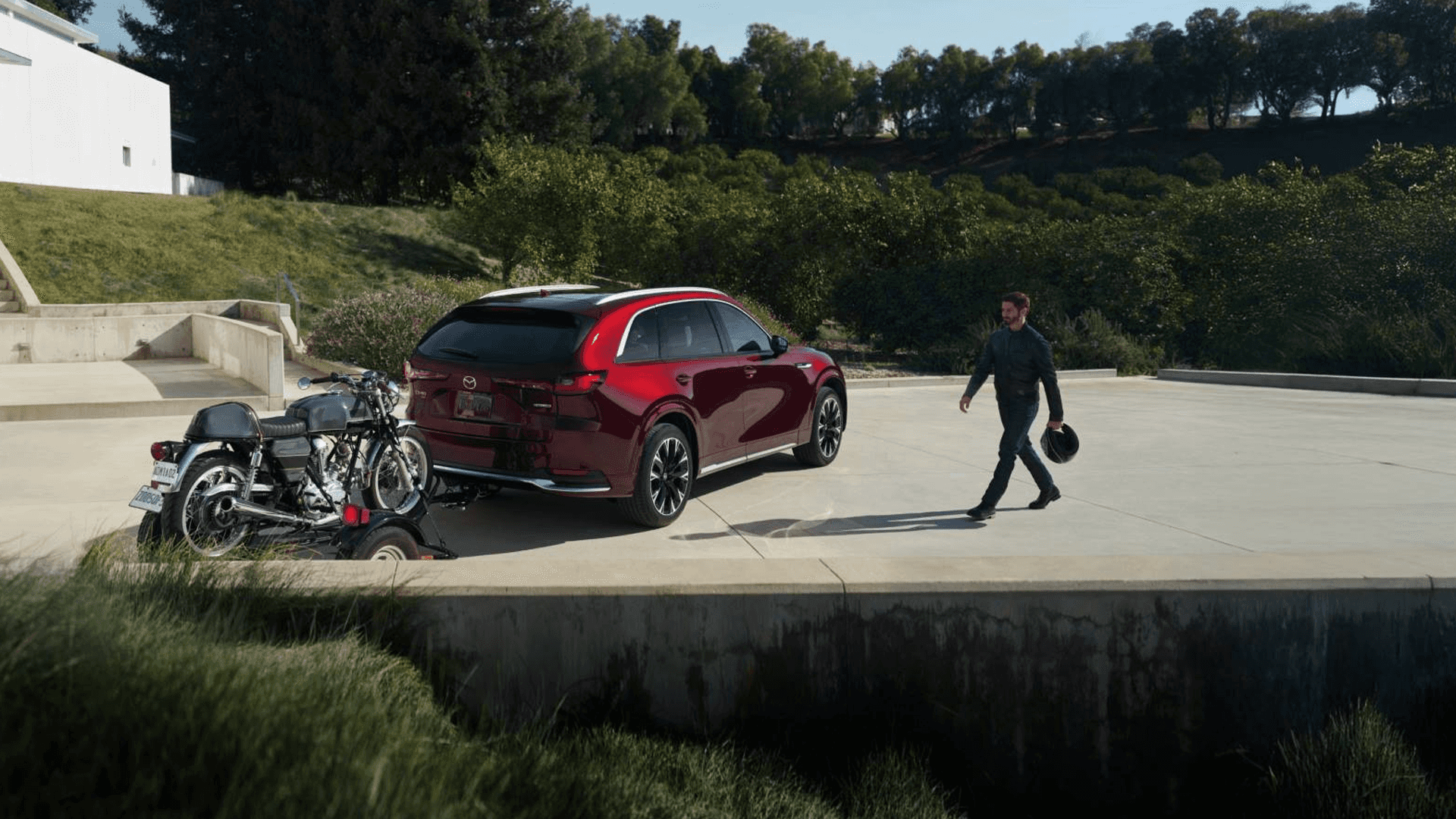 Red Mazda CX-90 parked near a trailer with a motorcycle as a person walks nearby in an outdoor setting