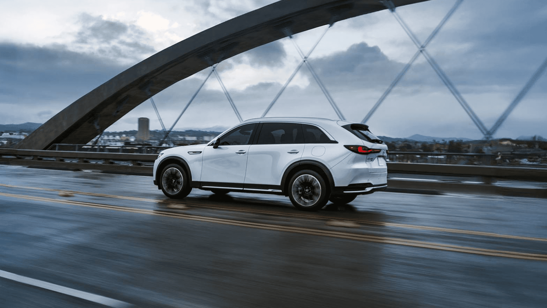 White Mazda CX-90 driving across a bridge on a wet road with city buildings and cloudy sky in the background