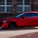 Red Mazda3 hatchback driving past a brick building, showing side profile, black wheels, and tinted windows