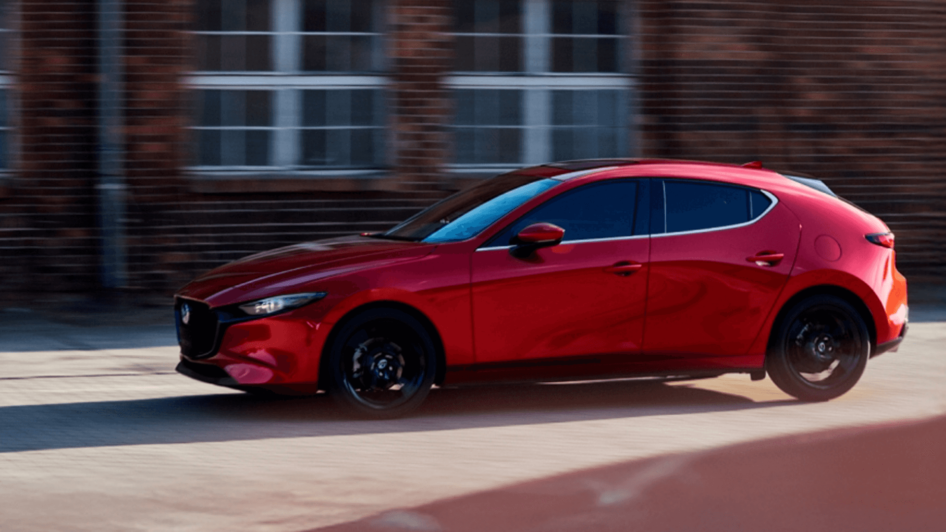 Red Mazda3 hatchback driving past a brick building, showing side profile, black wheels, and tinted windows