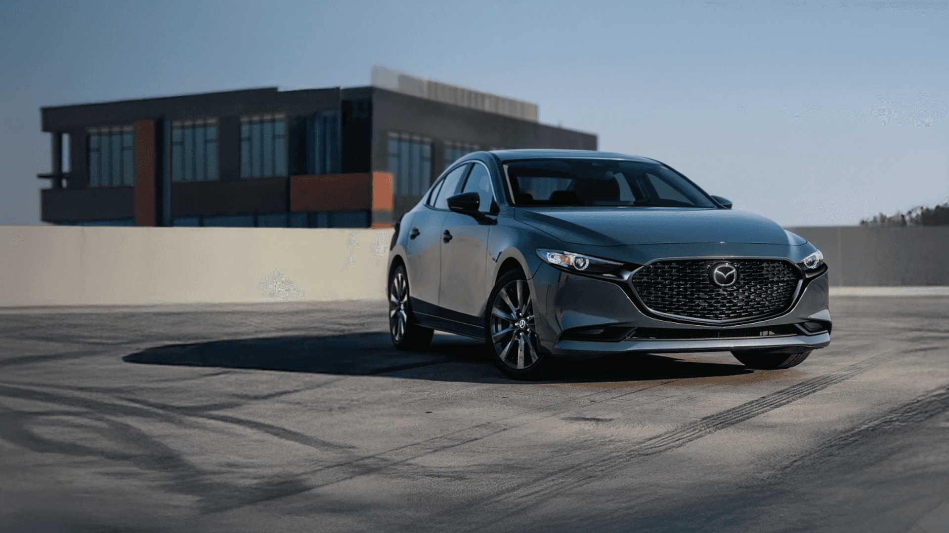 Gray Mazda3 sedan parked on pavement, showing front grille, headlights, alloy wheels, and sleek body lines