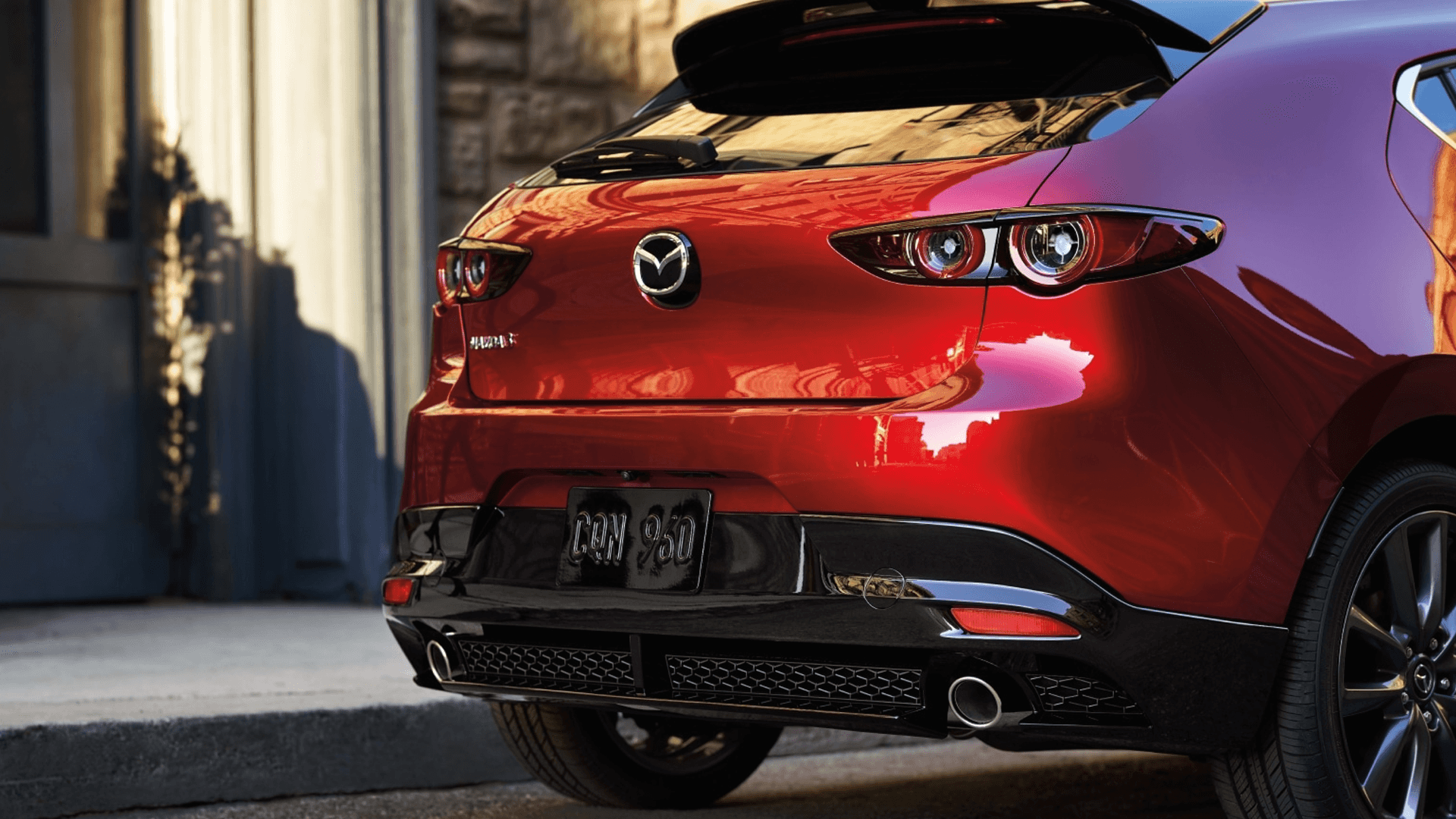Rear three-quarter view of a red Mazda3 hatchback with round taillights, roof spoiler, and dual exhaust outlets