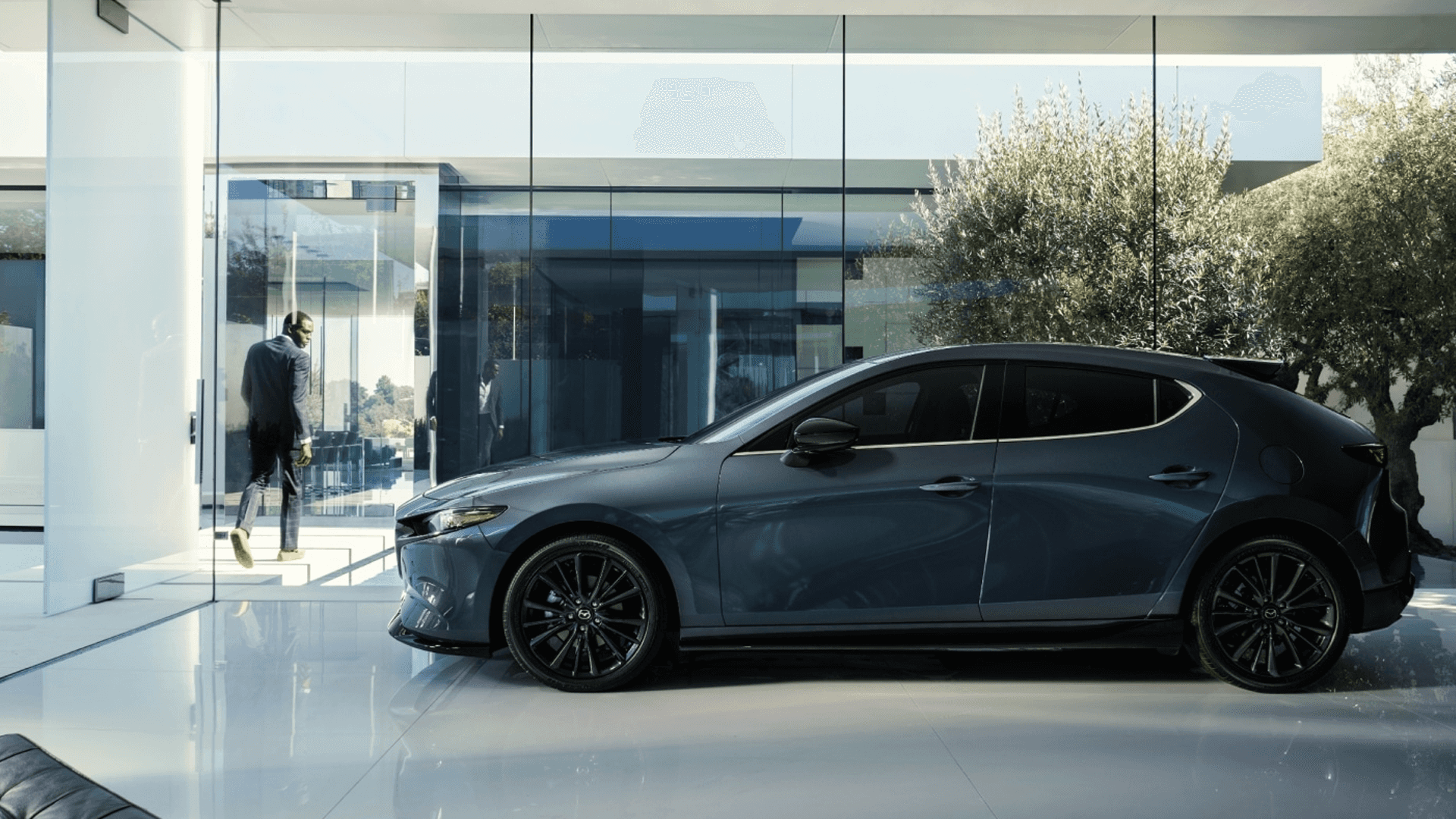 Gray Mazda3 hatchback in side profile indoors, showing black wheels, dark mirrors, and sculpted body sides