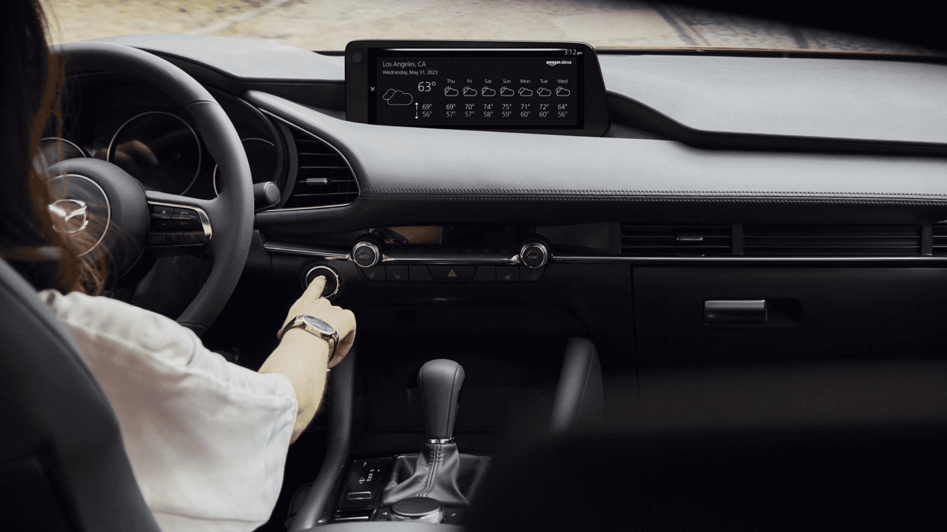 Dashboard with weather on center display, steering wheel, gear shifter, and hand pressing a control knob