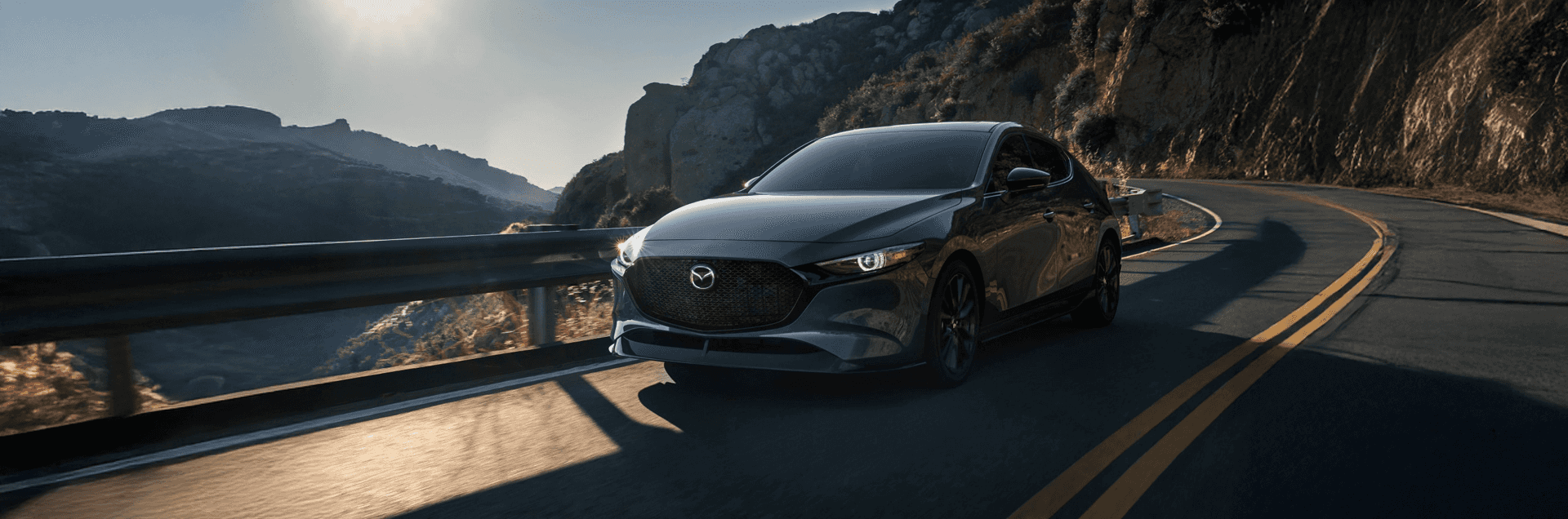 Dark hatchback driving on a winding mountain road beside a guardrail with sunlight over rocky hills