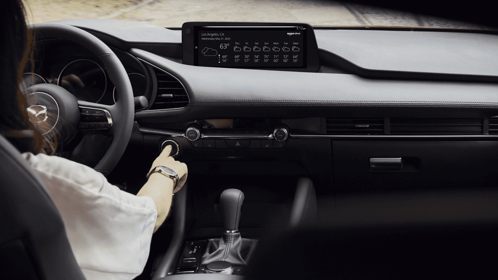 Driver reaching toward start button in Mazda3 interior with dashboard, center display, steering wheel, and gear selector visible
