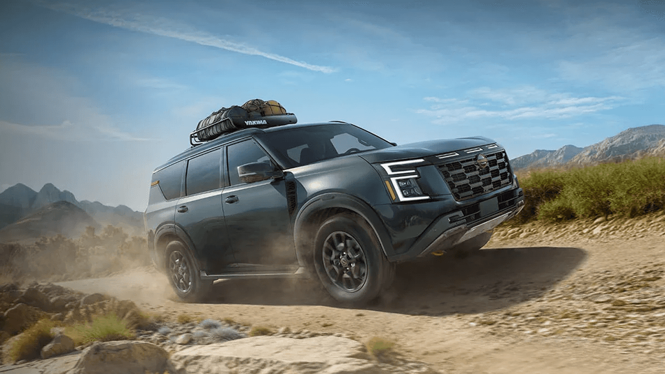 Green Nissan Armada driving on a dusty off-road trail with roof cargo box and mountains in the background