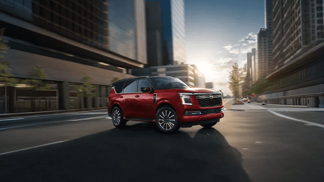 Red Nissan Armada driving through a city street with tall buildings and sunlight reflecting off glass facades