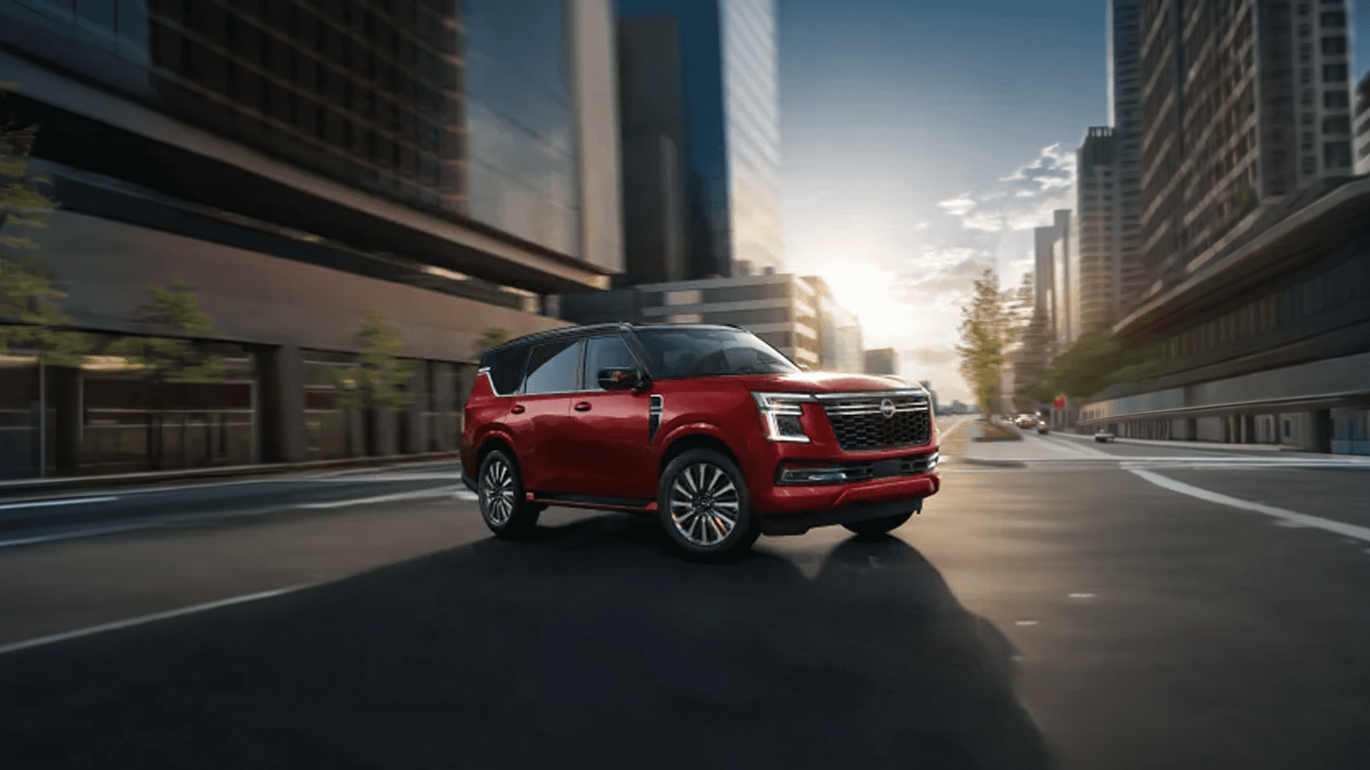 Red Nissan Armada driving through a modern city street with tall buildings and sunlight in the background