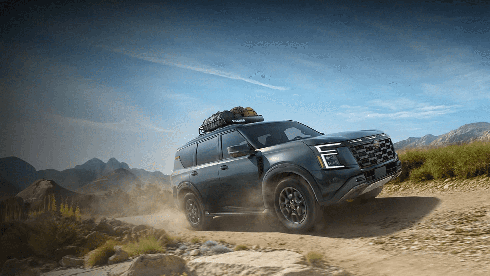 Green Nissan Armada driving on a dusty off-road trail with mountains and a roof rack carrying gear