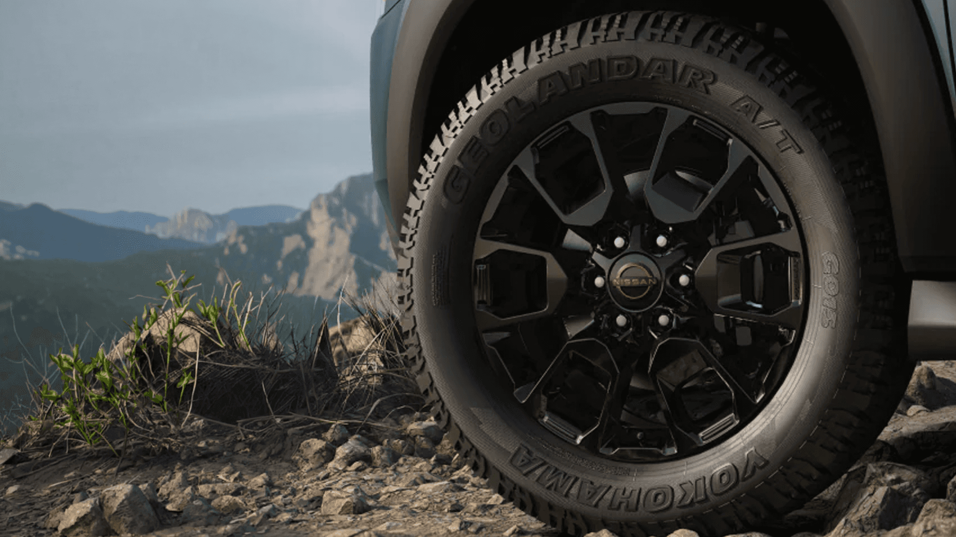 Close-up of a black alloy wheel with all-terrain tire on a rocky surface with mountains in the background
