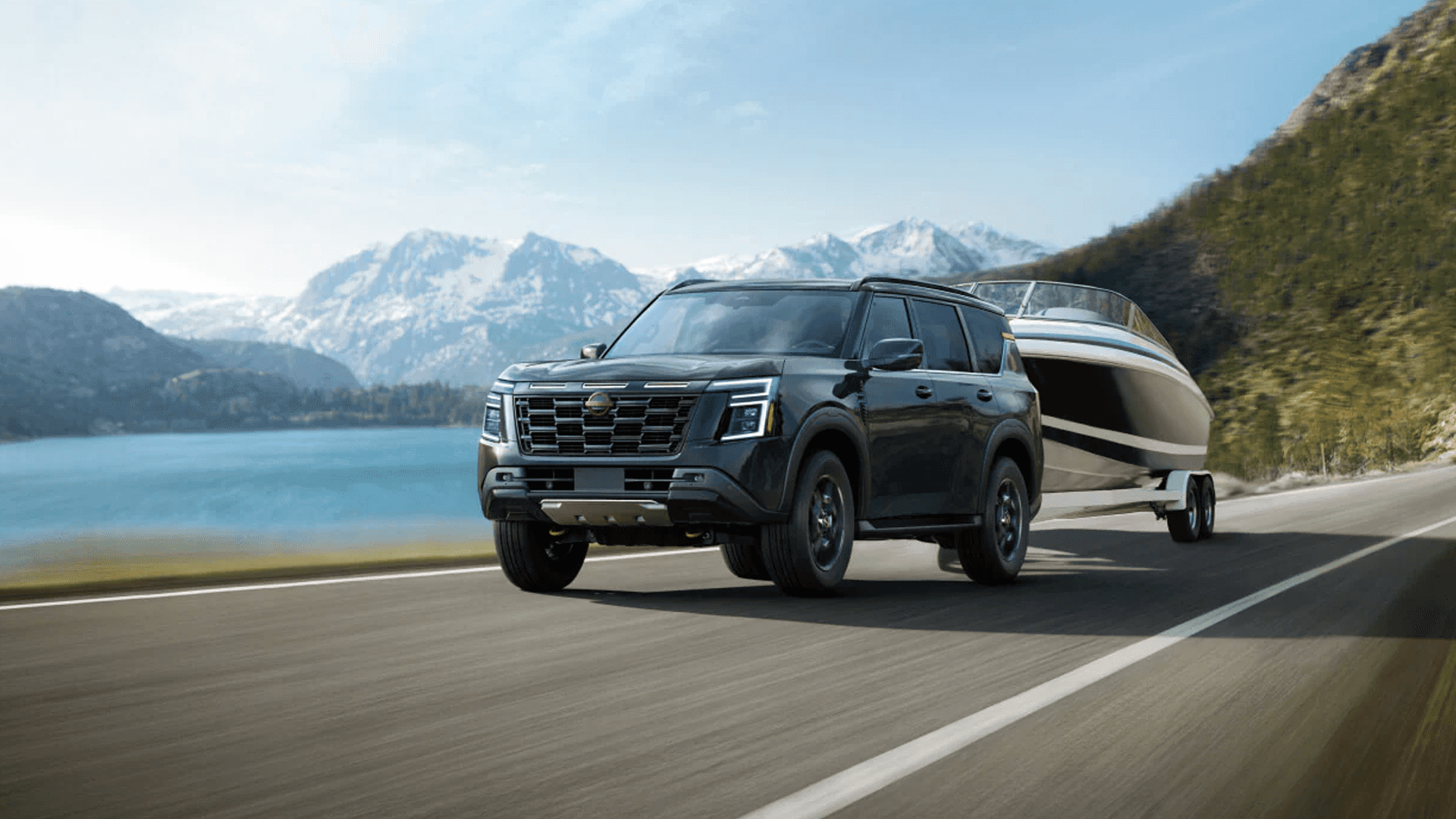 Black Nissan Armada towing a large boat on a paved road beside a lake with mountains in the distance