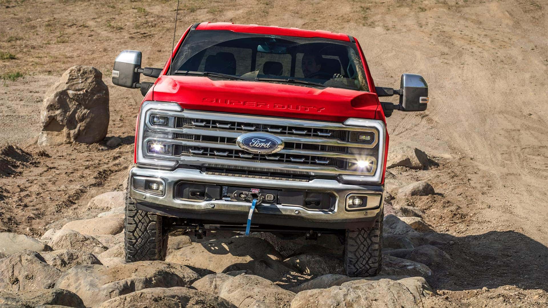 Front view of red 2026 Ford F-350 Super Duty on rocky terrain, showing bold grille, LED headlights, and Super Duty hood design
