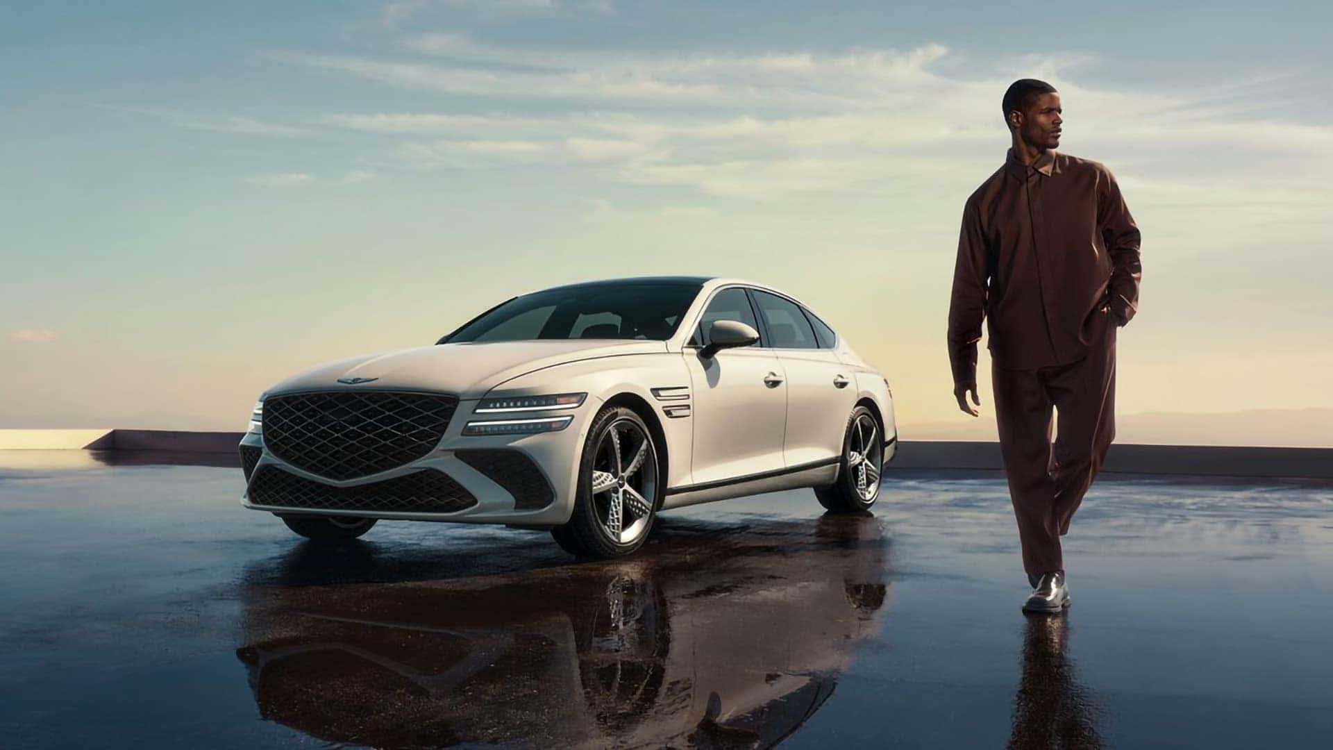 White 2026 Genesis G80 parked on reflective surface with driver walking nearby highlighting luxury sedan design