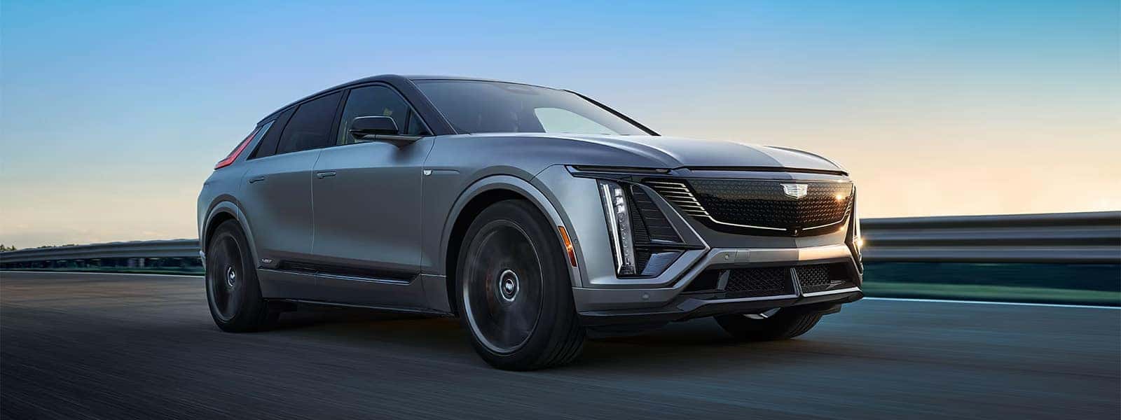 2026 Cadillac LYRIQ electric SUV driving on a highway at sunset with illuminated front lighting and Cadillac grille