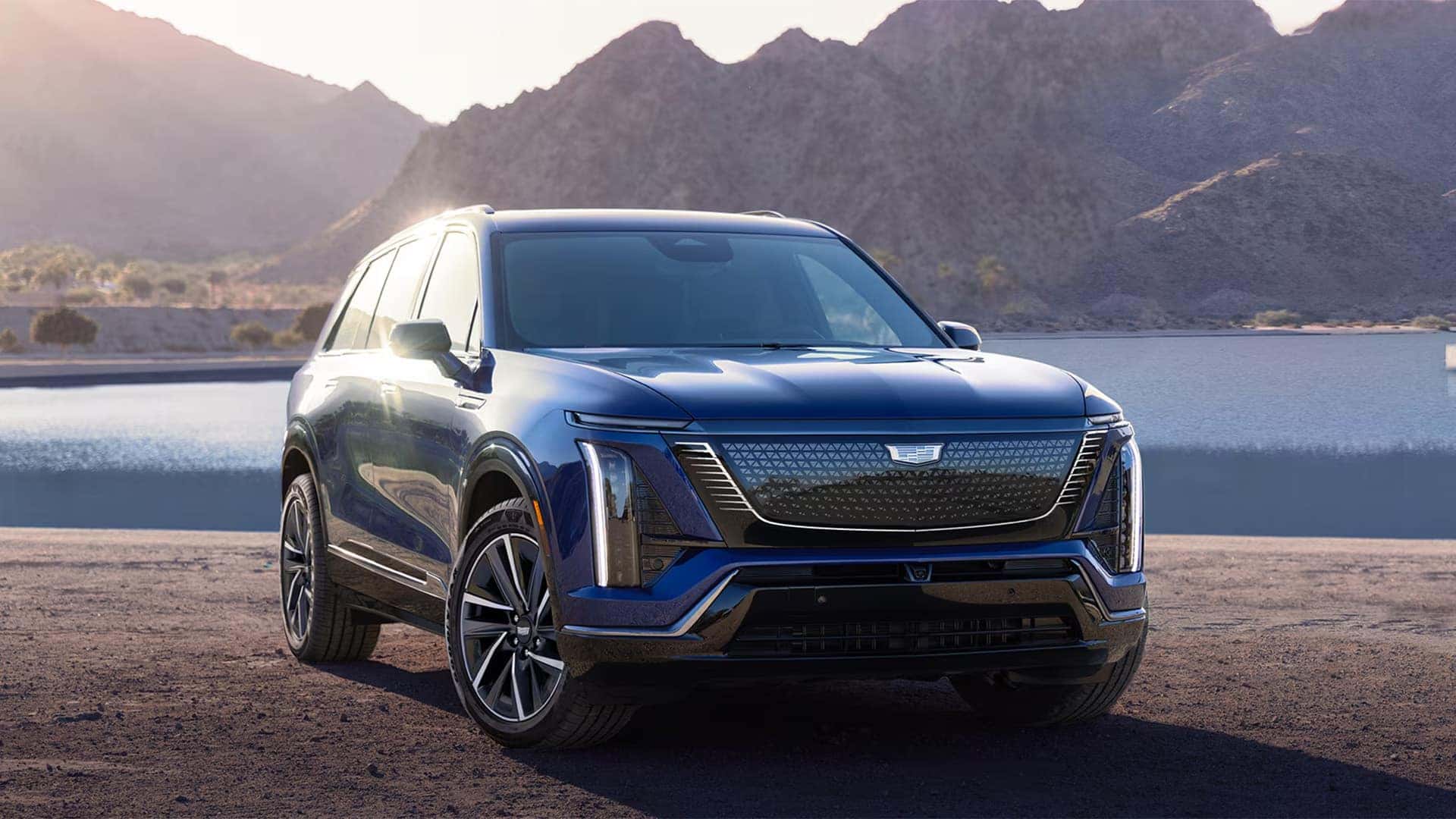 Blue 2026 Cadillac VISTIQ electric SUV parked near a mountain lake, showing the front grille, LED lighting, and alloy wheels