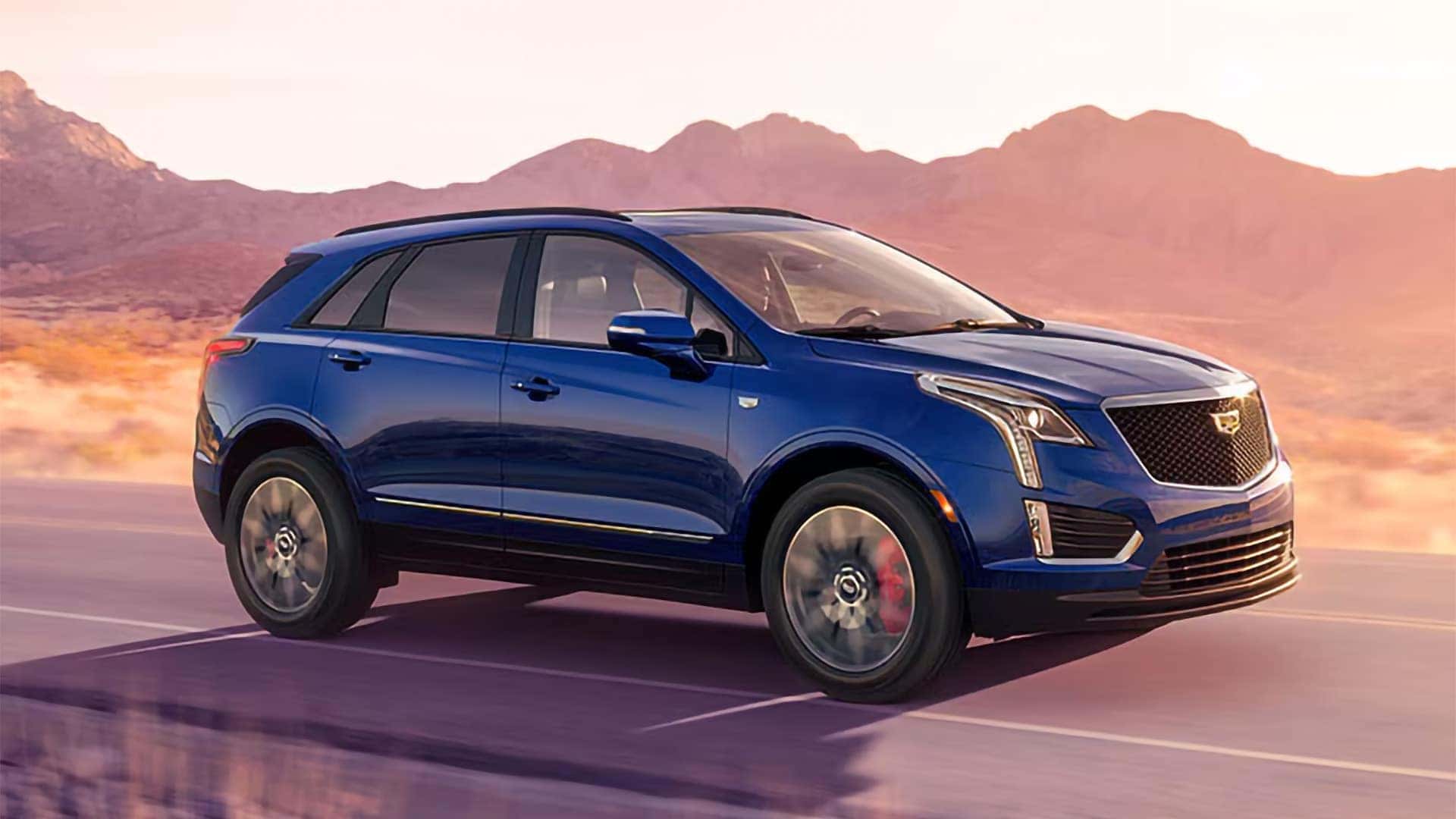 Blue 2026 Cadillac XT5 driving on a desert highway with chrome exterior accents, roof rails, and signature front lighting