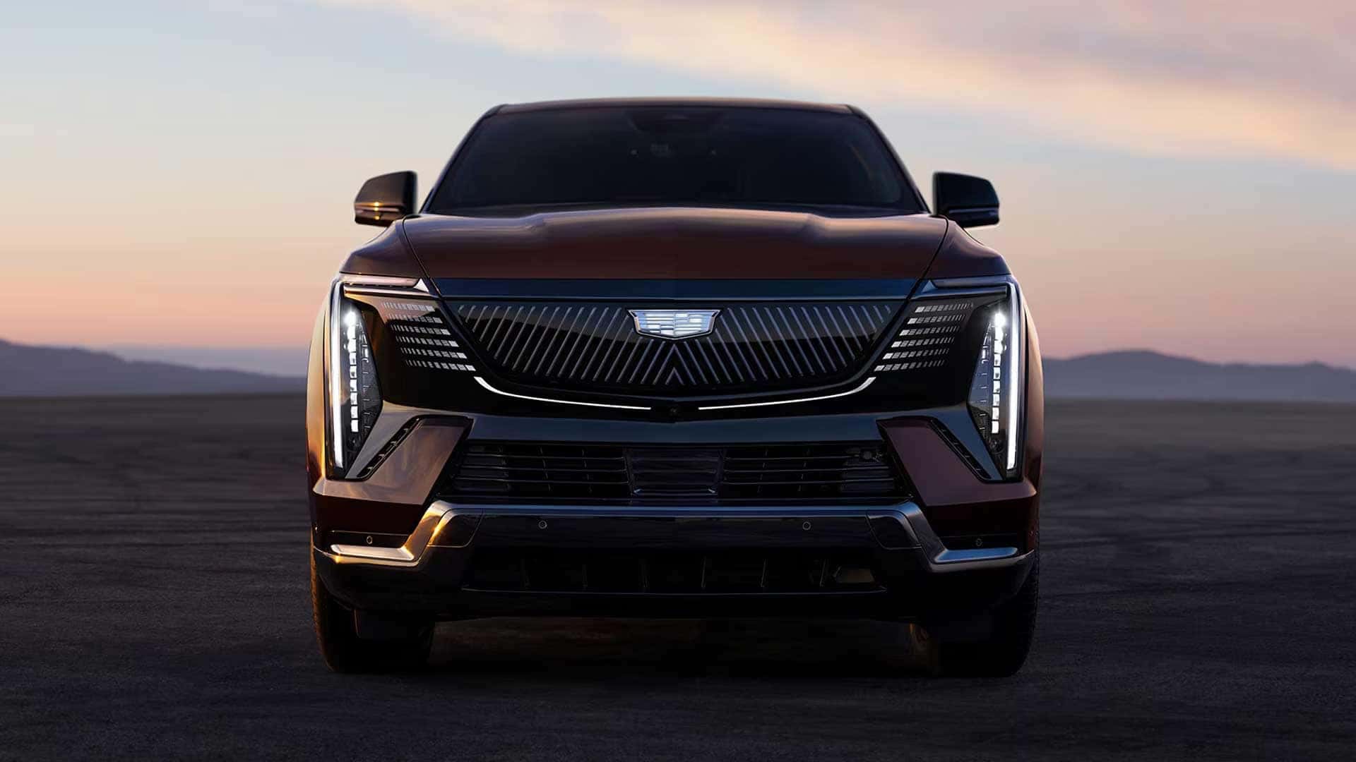 Front view of a red Cadillac Escalade IQ with illuminated vertical LED lights and grille, parked on open pavement at sunset