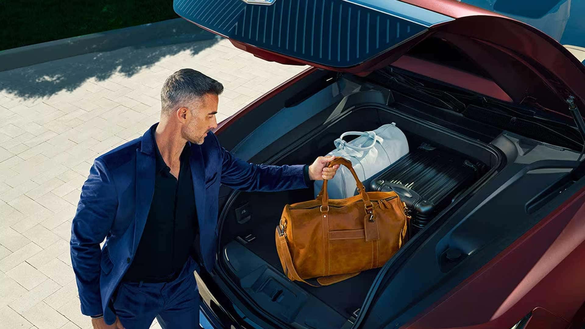 Man placing a brown leather bag into the front storage compartment of a red Cadillac Escalade IQ with open hood