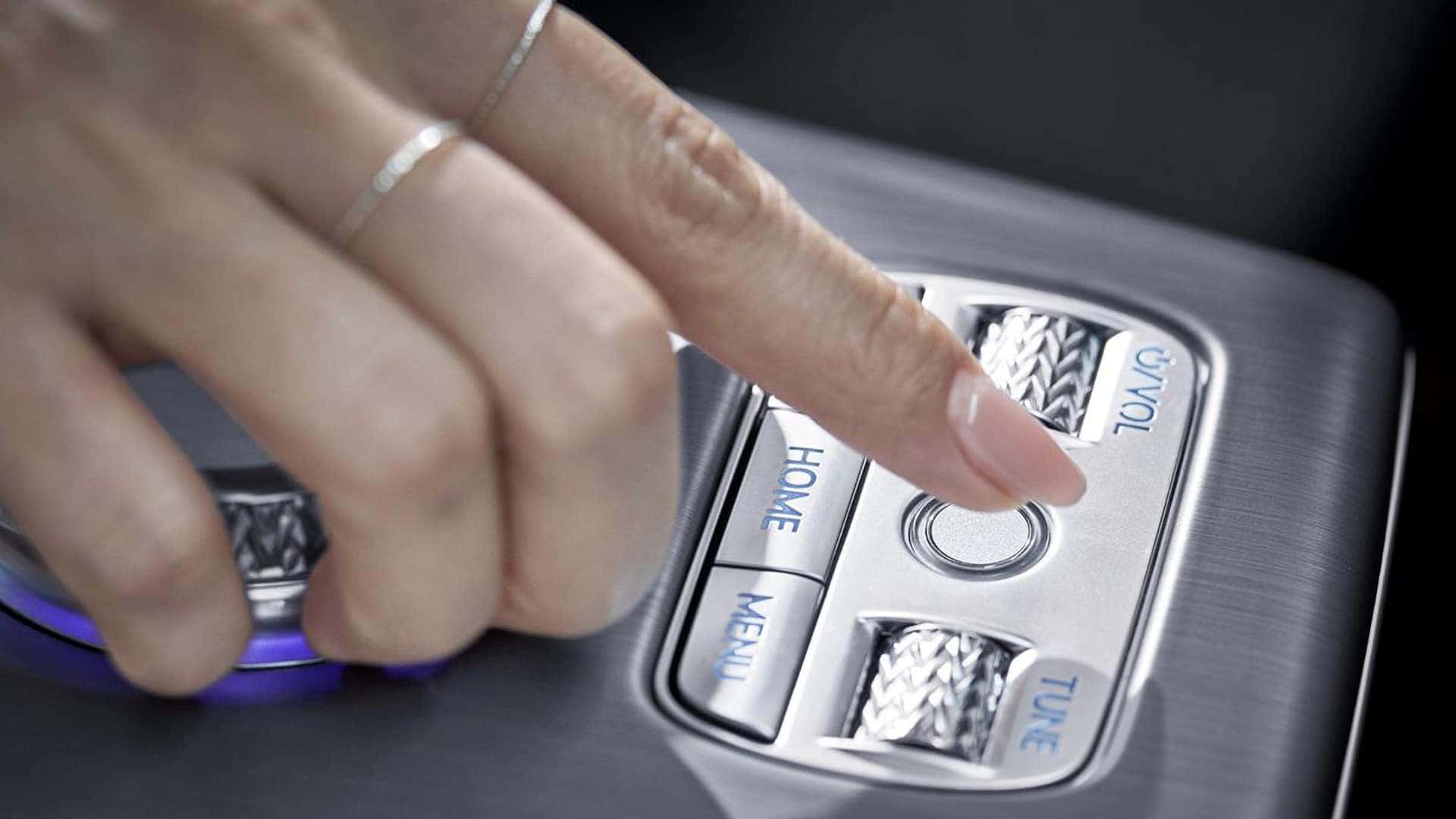 Driver using the 2026 Genesis GV60 fingerprint reader on the center console near home, menu, and tuning controls
