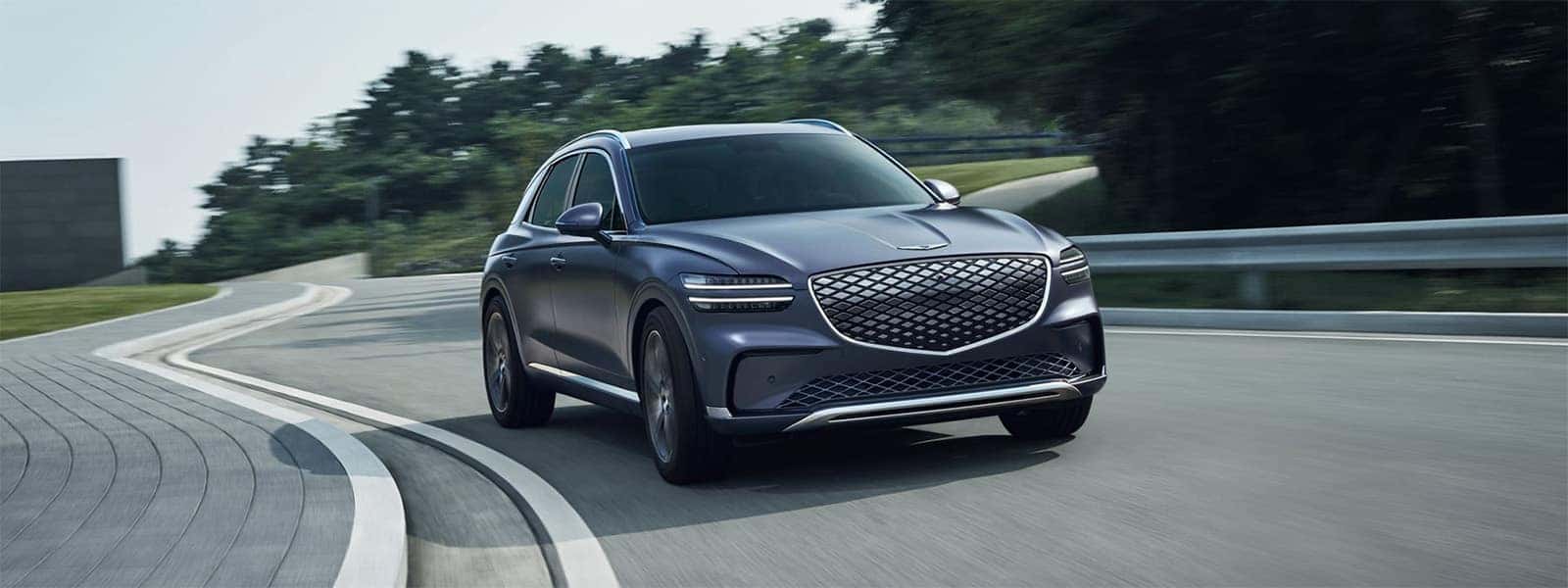Blue 2026 Genesis Electrified GV70 driving on a curved road, shown from the front three-quarter exterior view