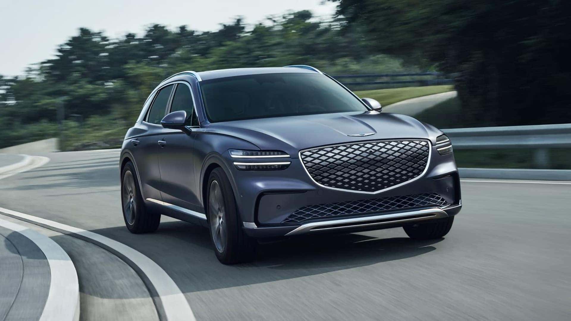 2026 Genesis Electrified GV70 driving on a curved road with front grille and LED lighting visible