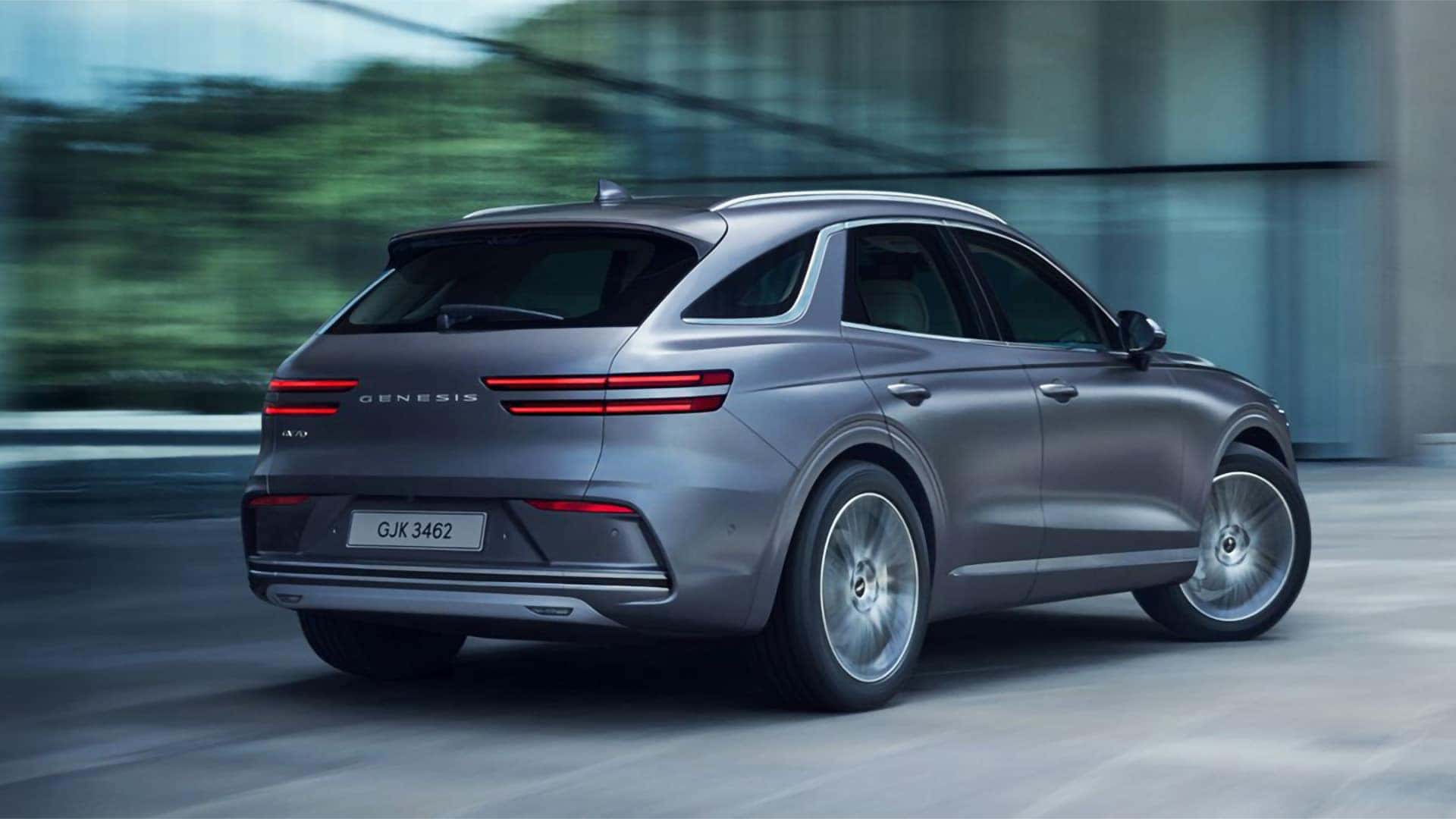 Gray 2026 Genesis Electrified GV70 driving past a modern building, shown from the rear three-quarter exterior view