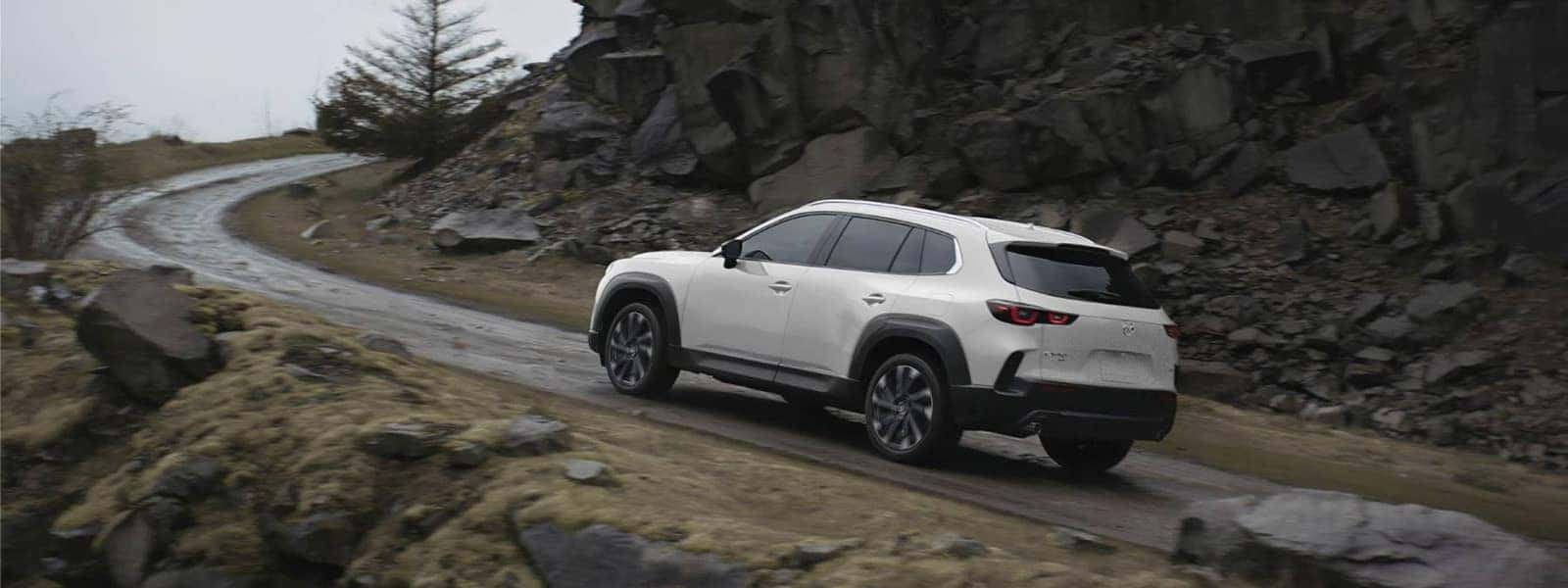 White 2026 Mazda CX-50 Hybrid driving uphill on a winding road through rocky terrain