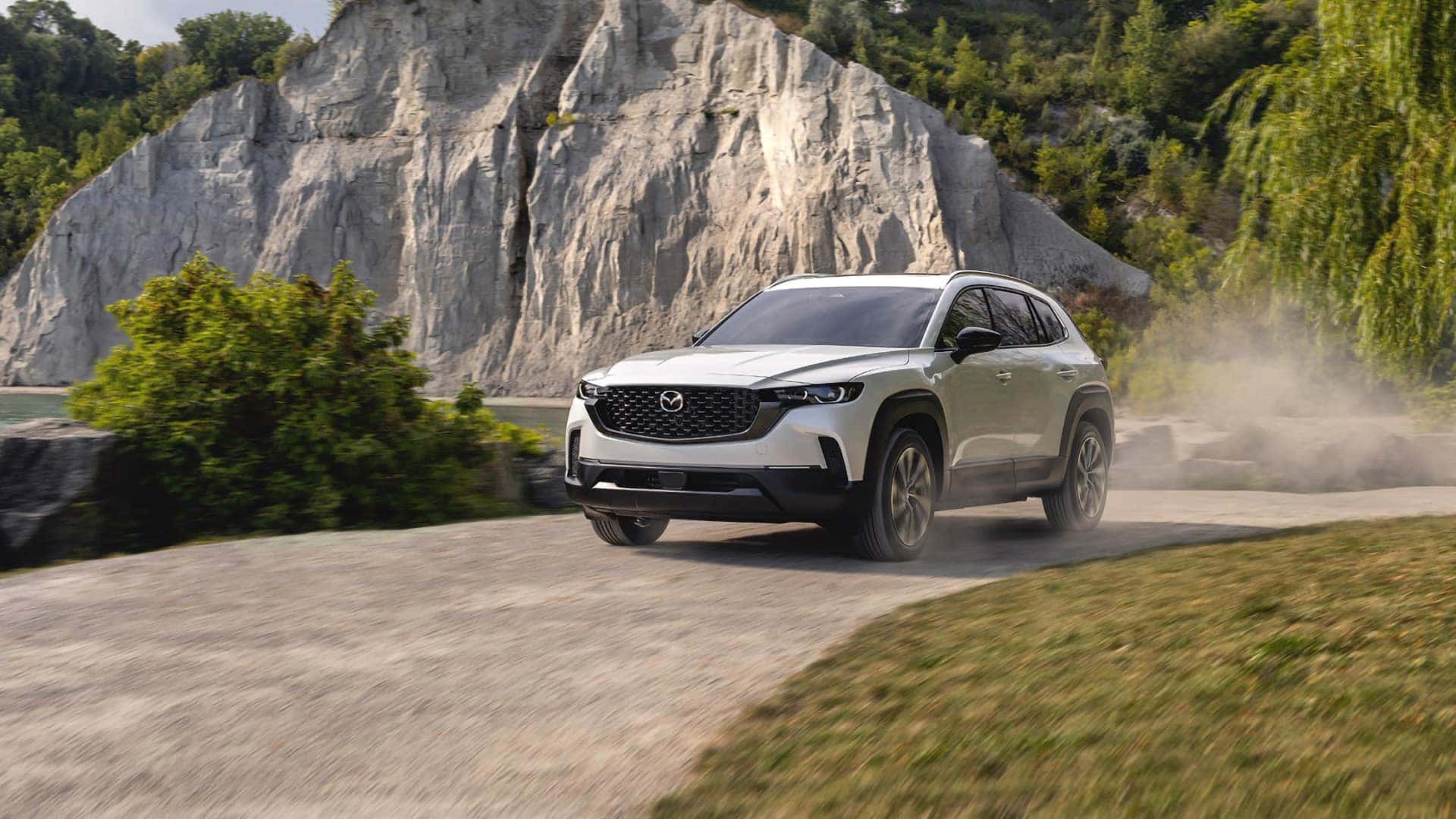 White 2026 Mazda CX-50 Hybrid driving past rocky cliffs, shown from the front three-quarter angle