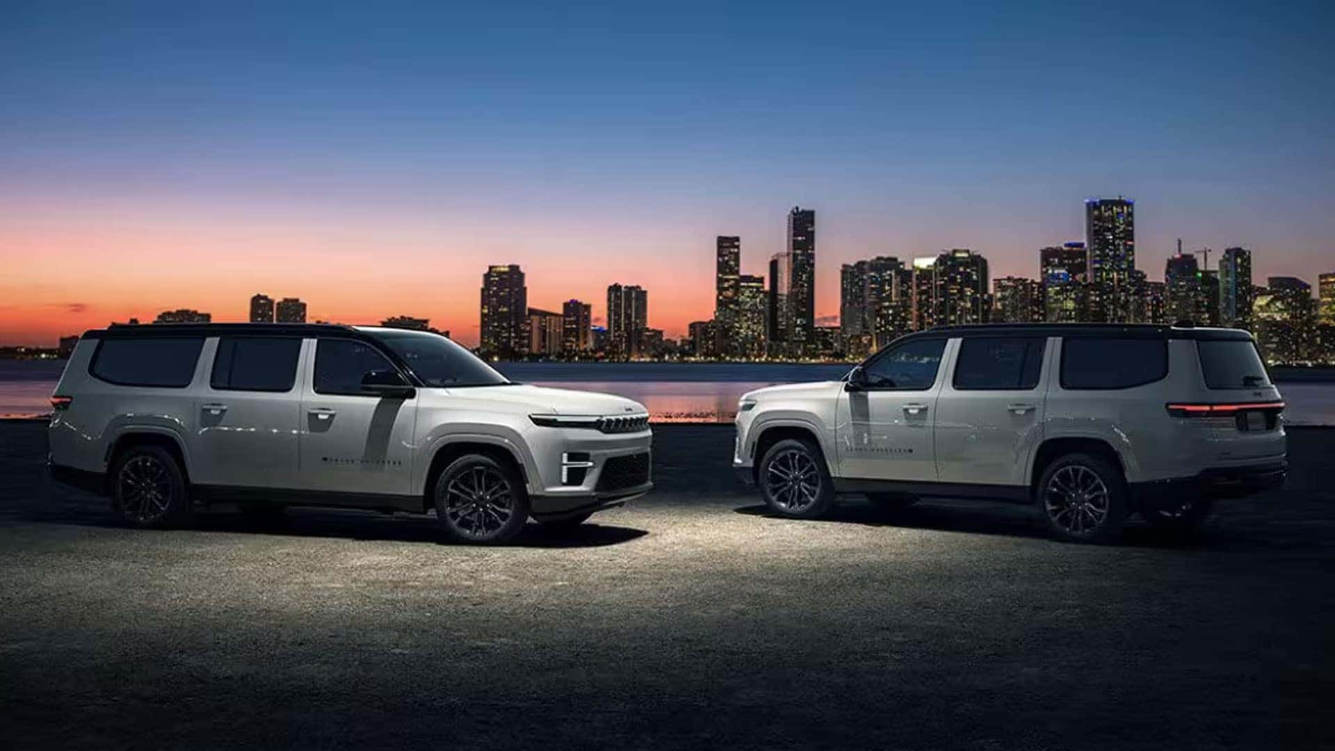 Two white Grand Wagoneer SUVs shown in side profile beside water with a city skyline at sunset.