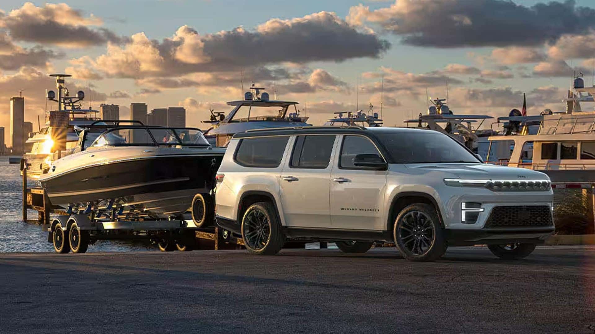 White Grand Wagoneer towing a black boat on a trailer beside a marina with boats and city buildings.
