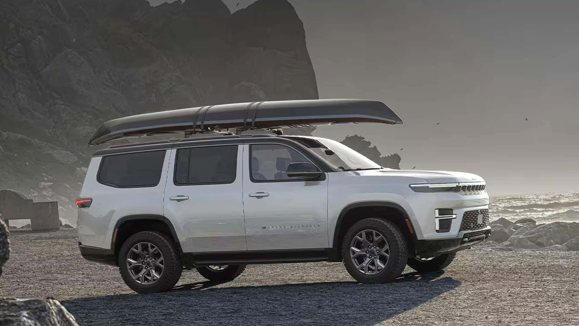White Grand Wagoneer in side profile on rocky coastal ground with a long canoe secured to the roof.