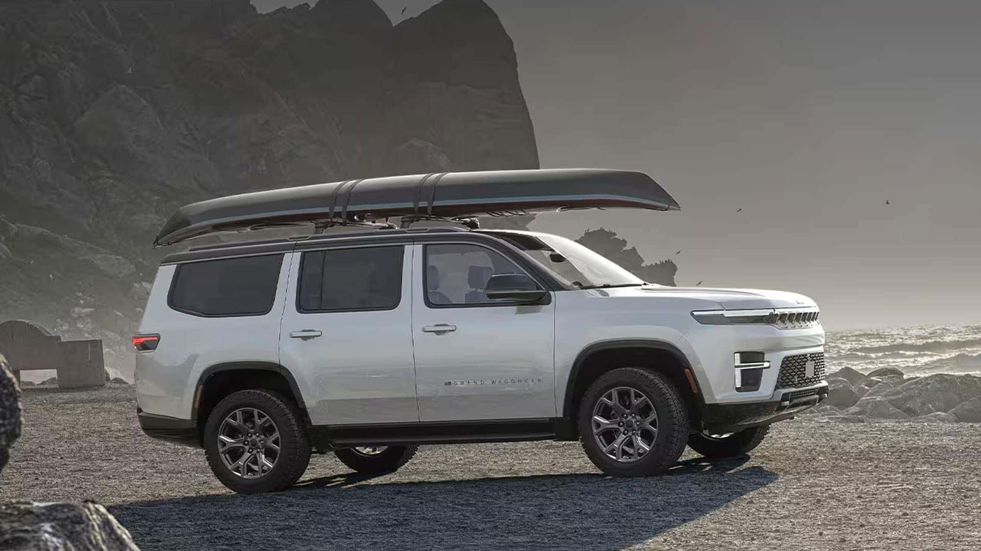 White 2026 Jeep Grand Wagoneer in side profile with a roof-mounted canoe, parked on rocky ground beside a coastal cliff.