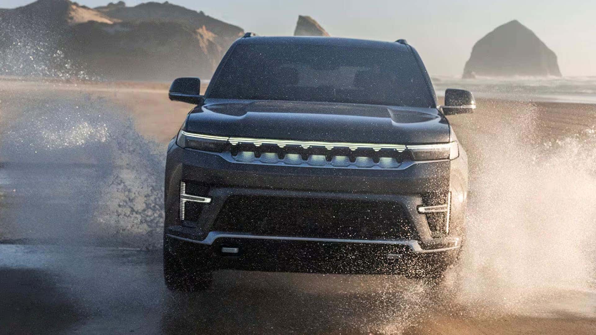 2026 Jeep Grand Wagoneer driving through shallow ocean water near a sandy beach and large rock formations.