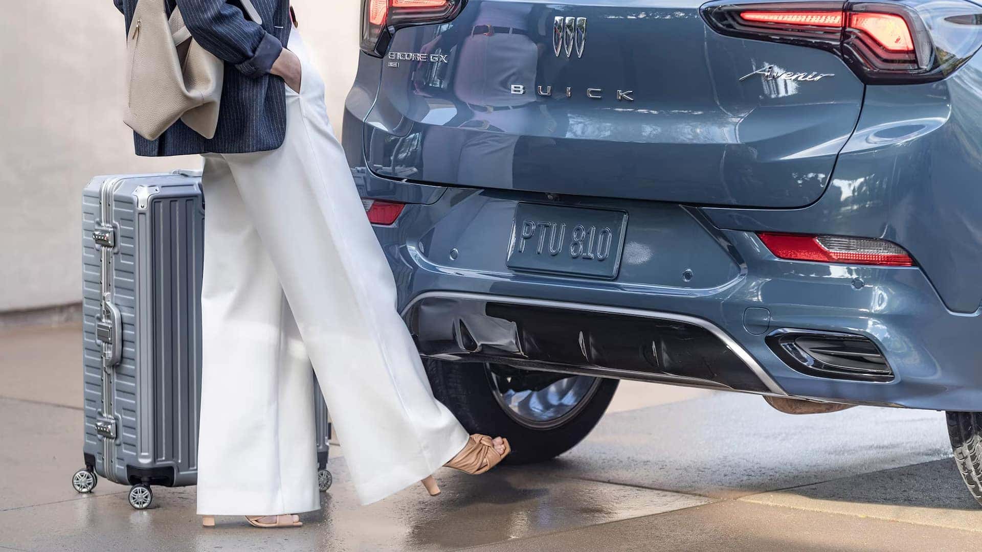 Woman using the hands-free power liftgate on a blue 2026 Buick Encore GX Avenir beside her luggage