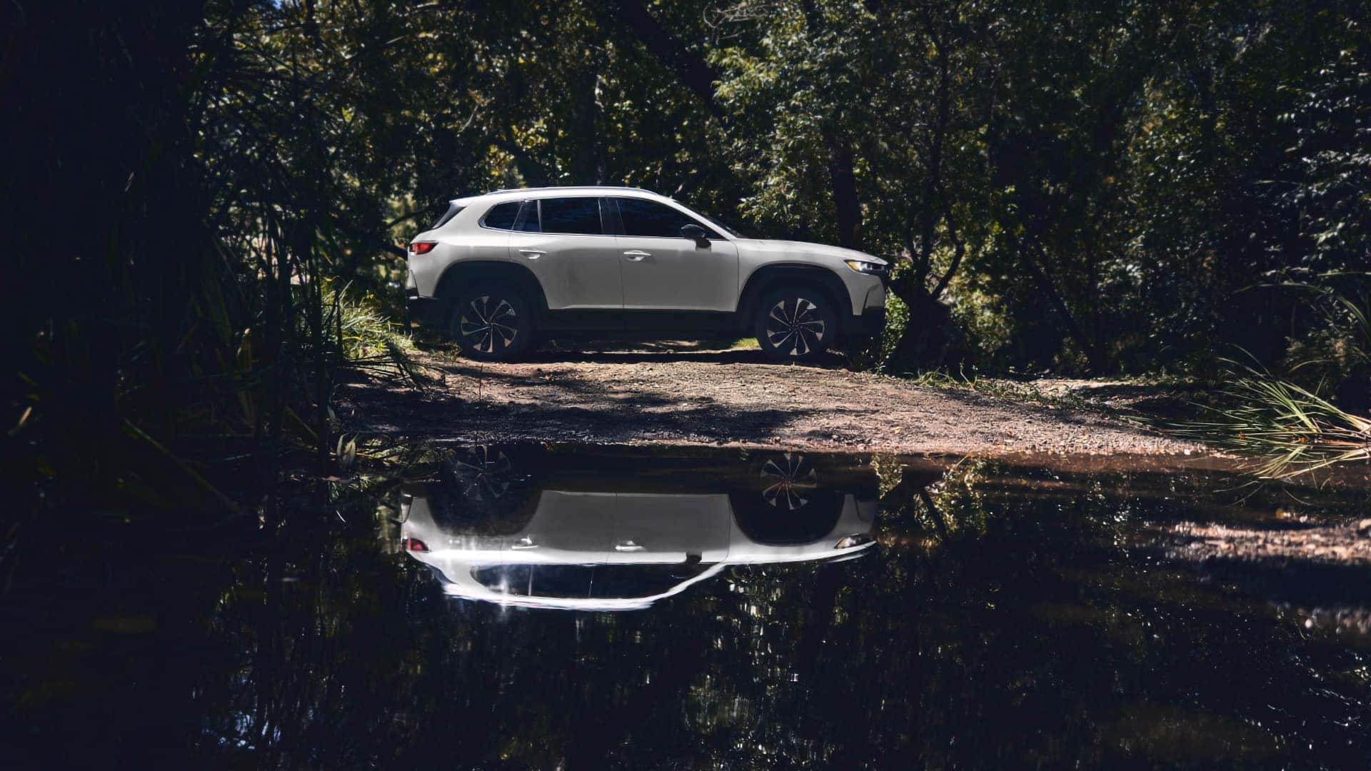 Side view of a white 2026 Mazda CX-50 Hybrid reflected in water along a wooded trail