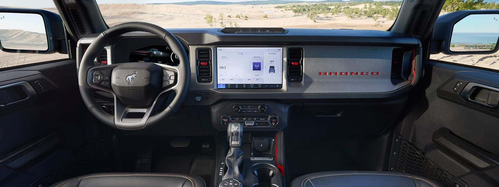 Ford Bronco interior with digital instrument cluster, center touchscreen, and terrain controls overlooking sand dunes