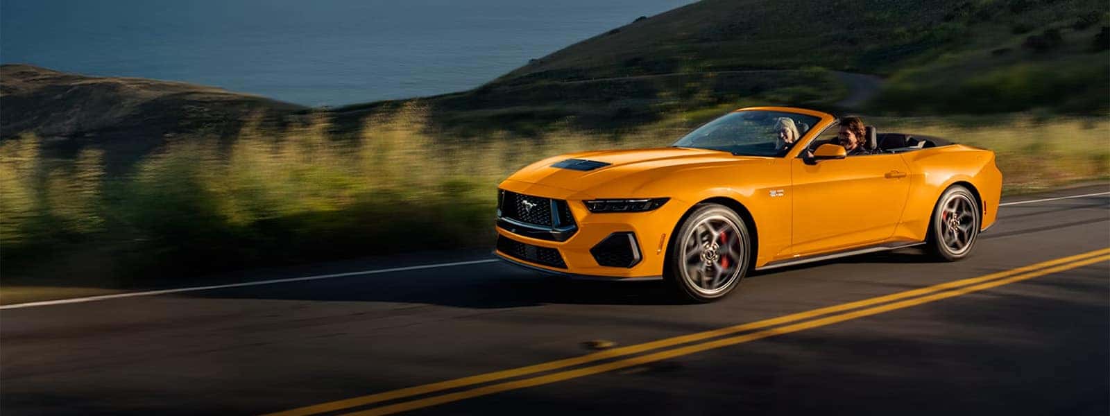 Orange 2026 Ford Mustang convertible driving on a winding road through green hills