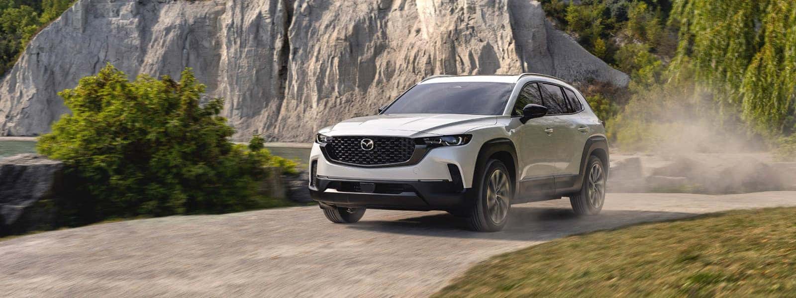 White 2026 Mazda CX-50 Hybrid driving on a gravel road beside rocky cliffs