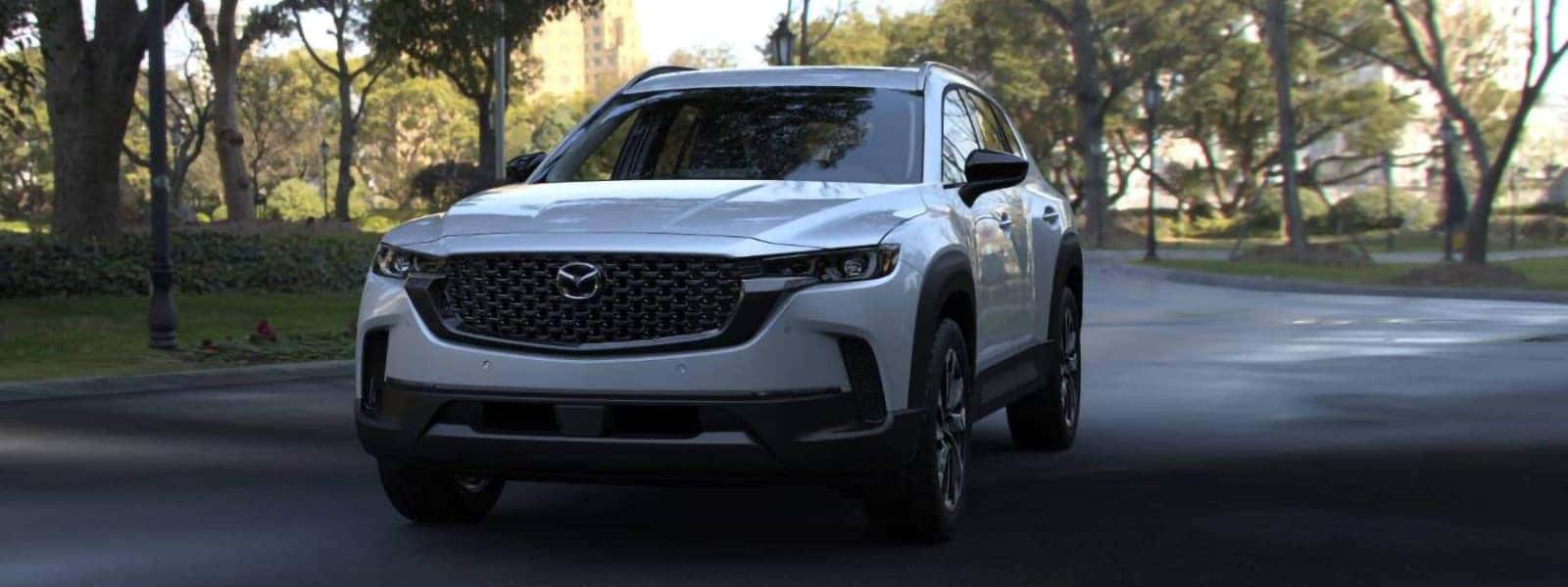 White 2026 Mazda CX-50 Hybrid driving on a tree-lined city road