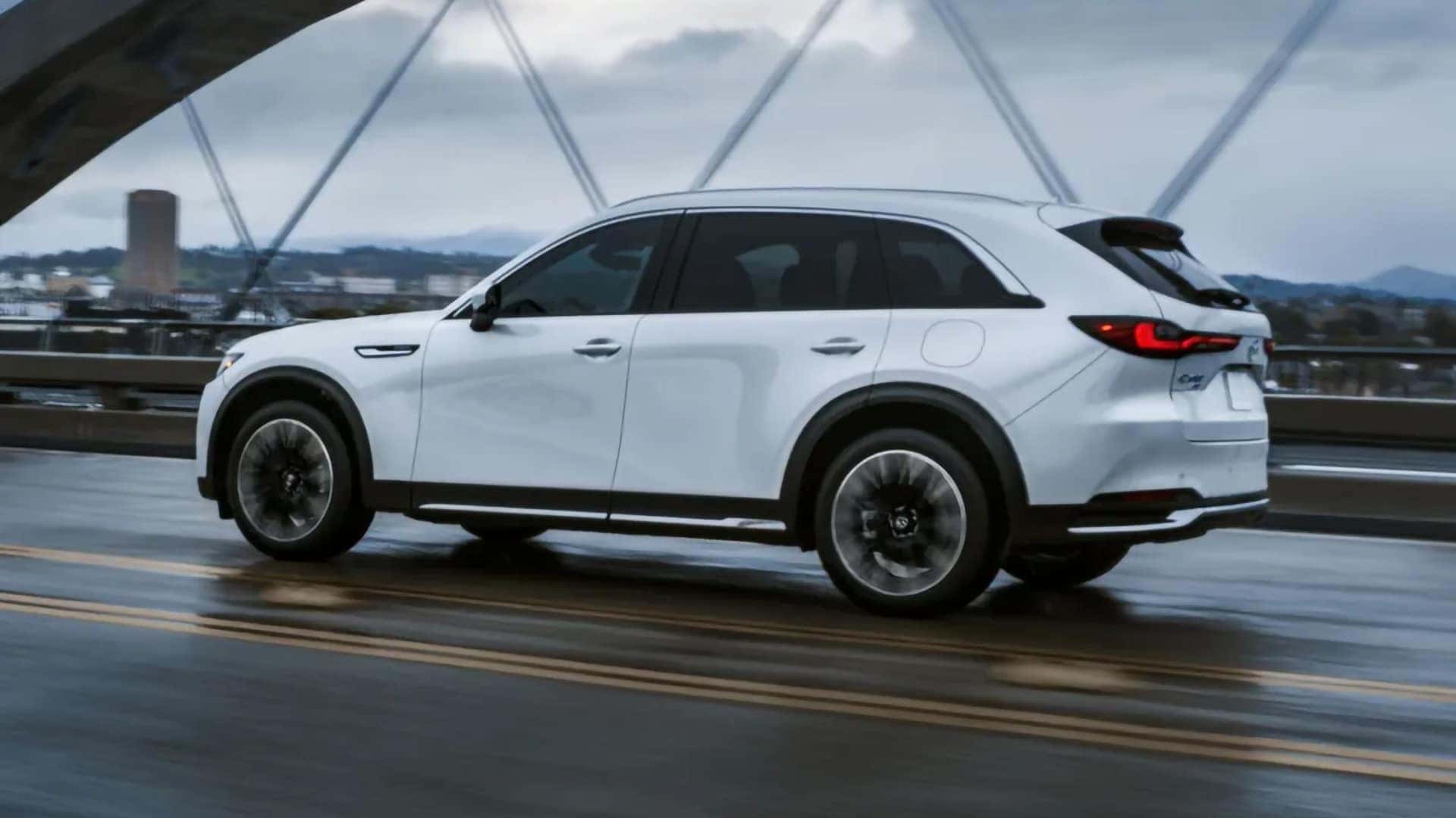 White 2026 Mazda CX-90 PHEV driving across a wet bridge, shown from the rear side angle