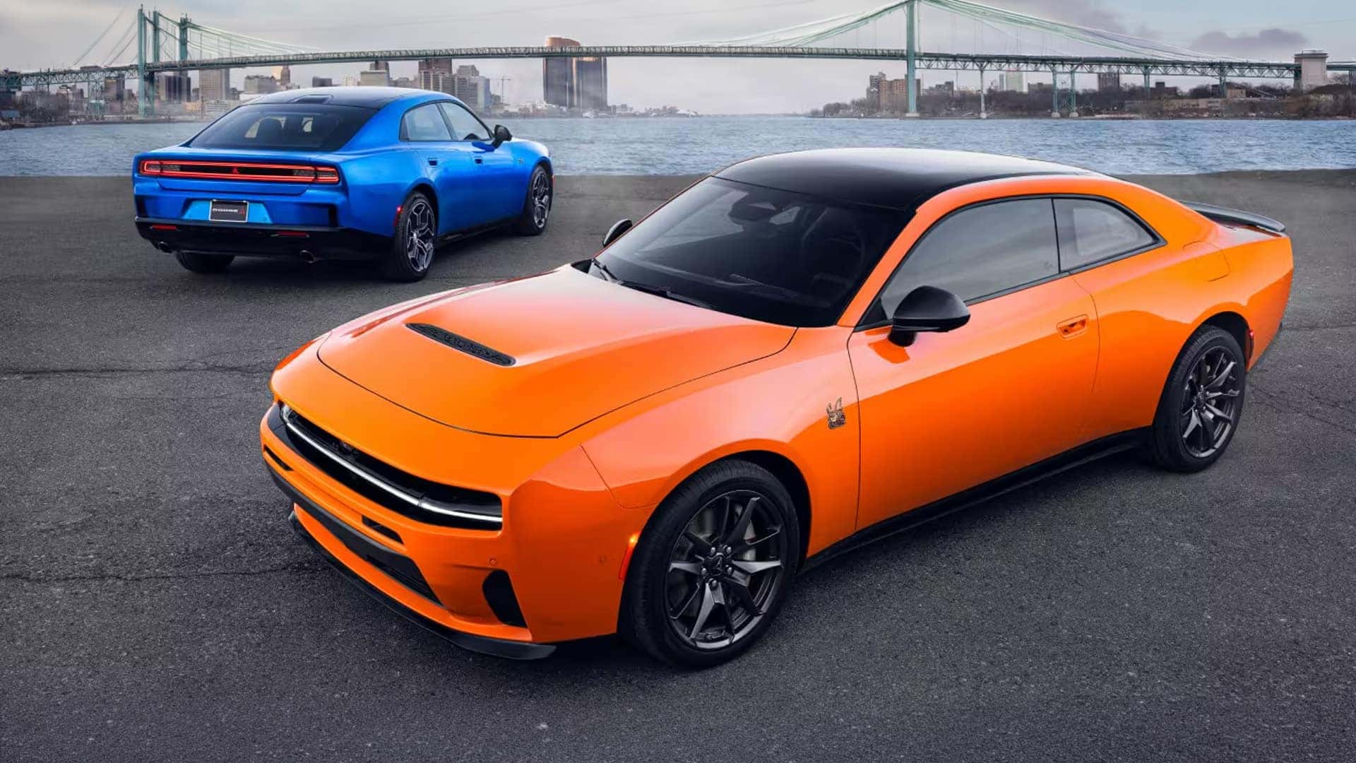 Orange two-door Dodge Charger in foreground and blue four-door Dodge Charger by water with suspension bridge behind.