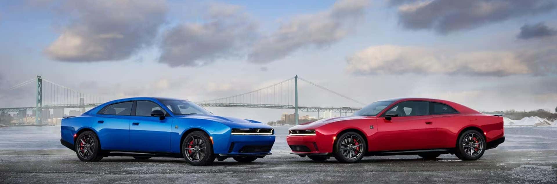 Blue four-door Dodge Charger and red two-door Dodge Charger parked by water with a suspension bridge behind.