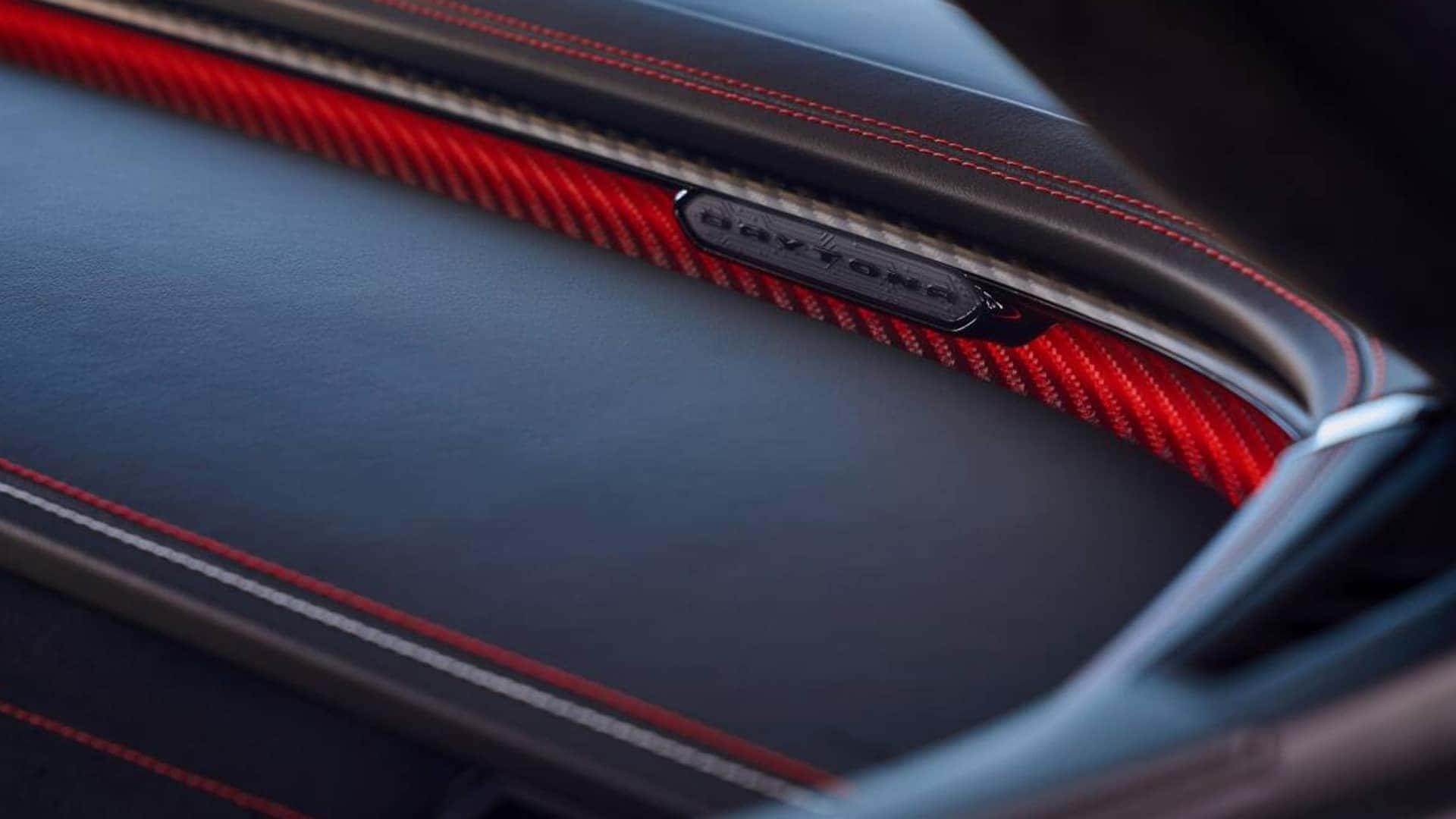 Close-up of black dashboard trim with red stitching, a red patterned accent panel, and a small Daytona badge.